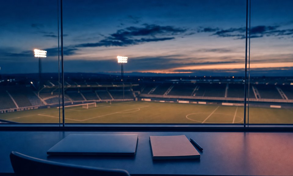 Lege perskamer achter glas in een stadion bij schemering, met een gesloten laptop en een notitieblok op het bureau