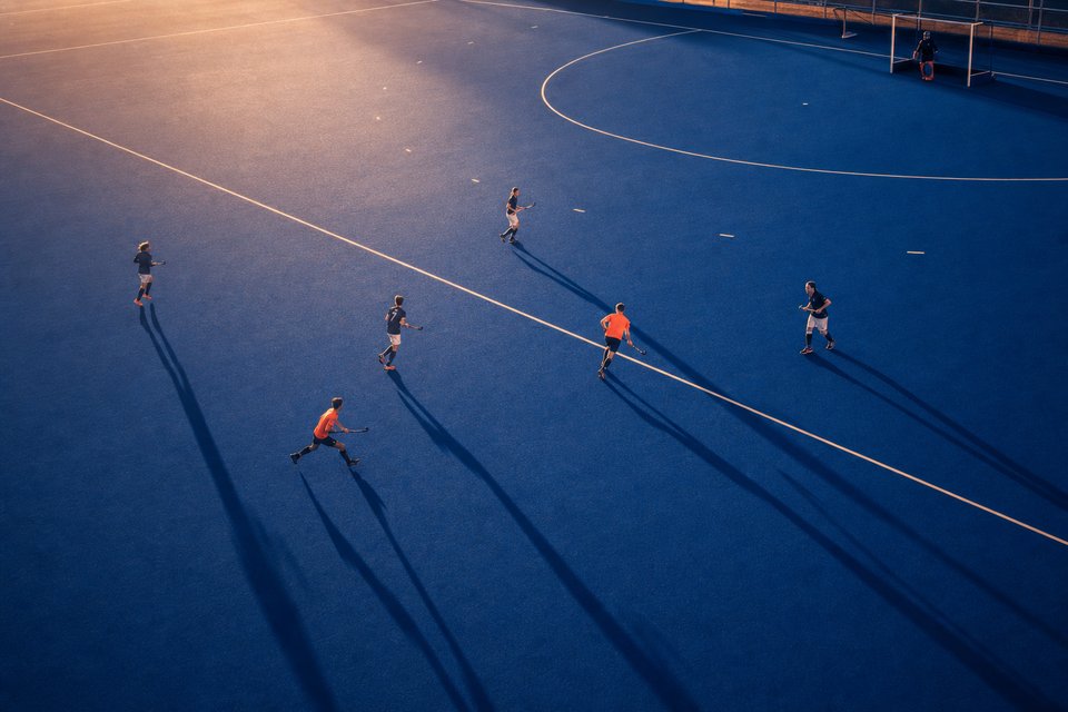 Blauw kunstgrasveld voor hockey in de late middagzon, van bovenaf gezien
